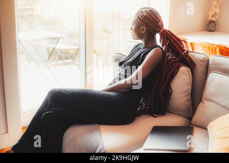 Eine Seitenansicht einer charmanten jungen schwarzen Frau, die zu Hause auf dem Sofa eines Gastzimmers sitzt und nachdenklich vor das Balkonfenster blickt, mit Stockfoto