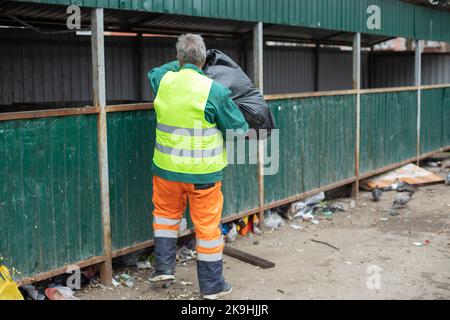 Mann wirft Müll in den Müll. Reiniger wirft Abfallbeutel aus. Werfen Sie einen schwarzen Beutel in den Tank. Arbeiter in heller Kleidung. Stockfoto