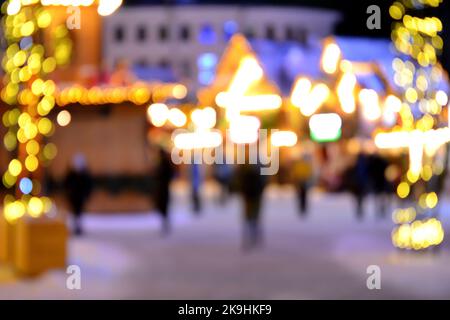 Unscharfer Hintergrund. In der Winternacht laufen die Menschen auf dem Stadtplatz. Schwarze Silhouetten von Menschen, die in der Nähe von Häusern gingen, schmückten leuchtende Beleuchtung. Weißlicht-Bokeh verwischt Punkte von glühenden Hauslichtern Stockfoto