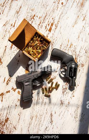 Eine halbautomatische Pistole von 9mm Glcck, ein Ruger 9 mm Revolver, Schalen und eine Schachtel Patronen in einer Pappschachtel auf einem Holztisch. Stockfoto