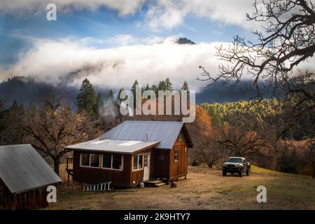 Holzhütte in den nebligen Bergen zur goldenen Stunde mit Rauch steigt aus der Stovepipe und LKW geparkt draußen Stockfoto