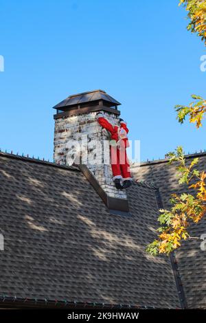 Entlümpelt Blow Up Santa auf Schornstein aussehen schlaff und wie er versucht zu hängen - lustig - mit Herbstblättern und Glühbirnen auf der Dachlinie - frühe Decora Stockfoto