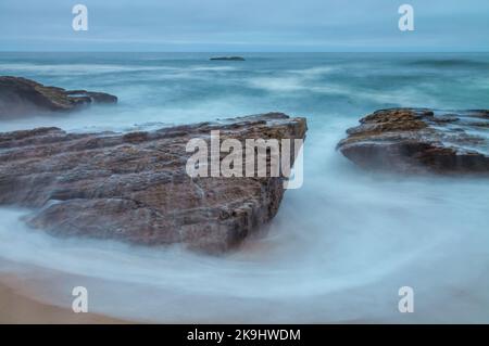 Ein Langzeitfoto erfasst die Bewegungen von Meereswellen, die auf felsige Felsvorsprünge an einem Strand stürzen. Stockfoto