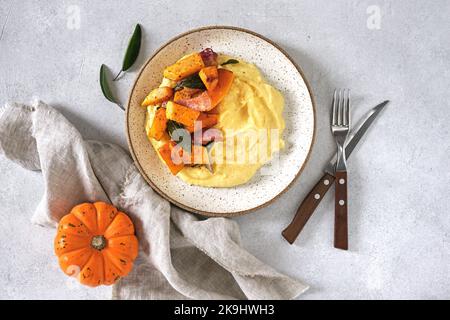 Polenta mit Kürbis, Salbei, becon in weißer Schale auf hellem Hintergrund. Traditionelle, einfache italienische Küche, vegane Küche. Draufsicht. Stockfoto