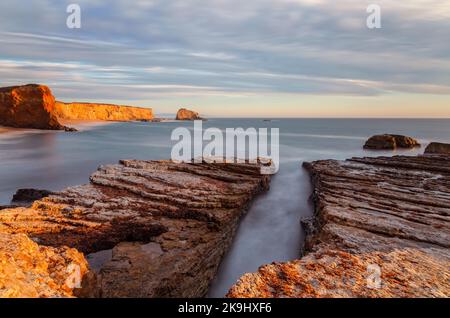 Eine atemberaubende Küstenszene bei Sonnenuntergang mit einer Reihe von geschichteten Felsformationen, durch die ein schmaler Wasserkanal fließt. Stockfoto