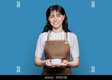 Junge lächelnde weibliche Kellnerin in Schürze mit Kaffeetasse, auf blauem Hintergrund Stockfoto