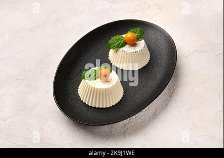 Hausgemachte zwei Panna Cotta mit natürlicher Vanilleschote, Physalis oder Winterkirsche und Minze auf schwarzem ovalem Teller Stockfoto