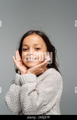 Aufgeregtes Kind in warmem Pullover, das die Hände in der Nähe des Gesichts hält und die Kamera isoliert auf Grau anschaut Stockfoto