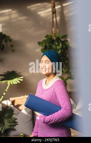 Glückliche birassische Frau, die Hijab trägt, Yogamatte hält und im Wohnzimmer lächelt Stockfoto