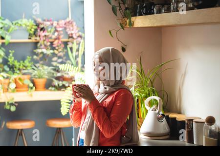 Glückliche birassische Frau, die Hijab trägt, in der Küche steht und Kaffee trinkt Stockfoto