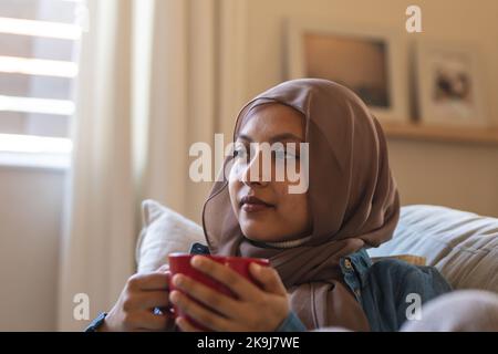 Glückliche birassische Frau, die Hijab trägt, auf dem Sofa im Wohnzimmer sitzt, Kaffee trinkt Stockfoto