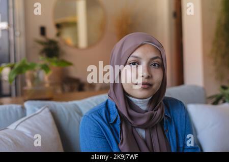 Porträt einer glücklichen, birassischen Frau, die Hijab trägt und auf dem Sofa sitzt Stockfoto