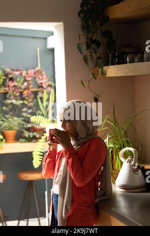 Glückliche birassische Frau, die Hijab trägt, in der Küche steht und Kaffee trinkt Stockfoto