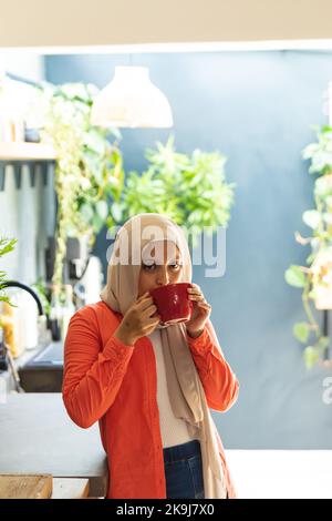 Glückliche birassische Frau, die Hijab trägt, in der Küche steht und Kaffee trinkt Stockfoto