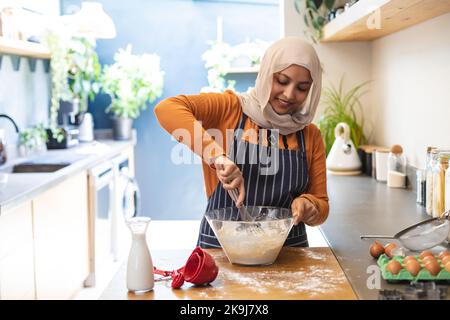 Glückliche birassische Frau, die Hijab trägt, in der Küche steht und backt Stockfoto