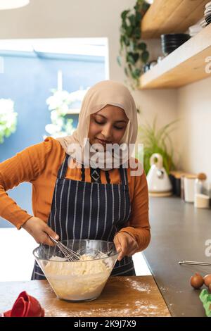 Glückliche birassische Frau, die Hijab trägt, in der Küche steht und backt Stockfoto