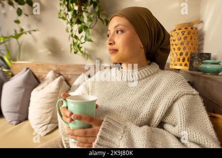 Glückliche birassische Frau, die Hijab trägt, auf dem Sofa im Wohnzimmer sitzt, Kaffee trinkt Stockfoto