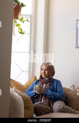 Glückliche birassische Frau, die Hijab trägt, auf dem Sofa im Wohnzimmer sitzt, Kaffee trinkt Stockfoto