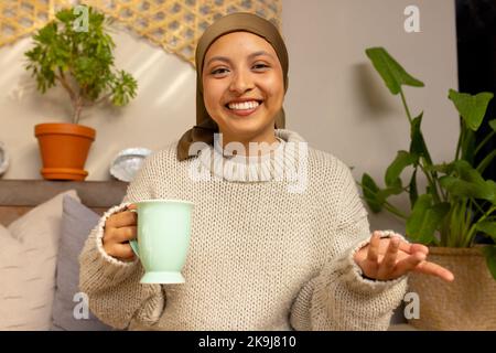 Glückliche birassische Frau, die Hijab trägt, ein Videogespräch hat, Kaffee trinkt Stockfoto