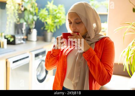 Glückliche birassische Frau, die Hijab trägt, in der Küche steht und Kaffee trinkt Stockfoto