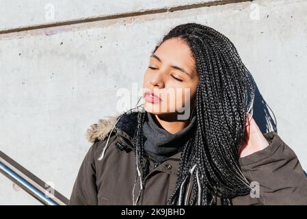 Ruhige Hündin mit Zöpfen, die schwarzes Haar berühren und sonniges Wetter mit geschlossenen Augen auf der Straße genießen Stockfoto