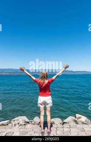 Blick von hinten auf eine blonde, nicht erkennbare Touristin mit erhobenen Armen vor dem Genfer See. Stockfoto