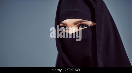 Es liegt in ihren Augen. Studioaufnahme einer jungen arabischen Frau, die eine Burka vor grauem Hintergrund trägt. Stockfoto