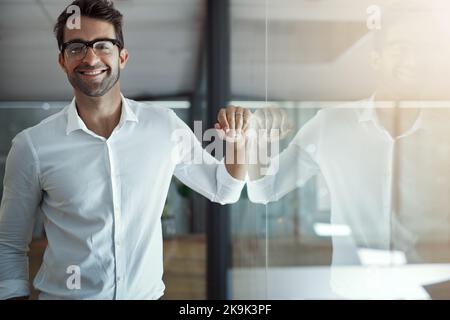 Ich habe es bis an die Spitze geschafft. Ein Porträt eines hübschen jungen Geschäftsmanns, der sich in seinem modernen Büro an einer Glaswand lehnt. Stockfoto