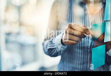 Einige gewinnbringende Ideen festhalten. Nahaufnahme einer unkenntlichen Geschäftsfrau, die in einem Büro Haftnotizen auf eine Glaswand klebte. Stockfoto