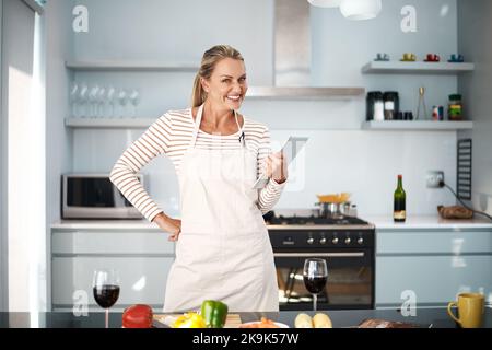 Ich freue mich, dieses neue Rezept auszuprobieren. Eine reife Frau benutzt ihr digitales Tablet, während sie ihre Schürze in der Küche trägt. Stockfoto