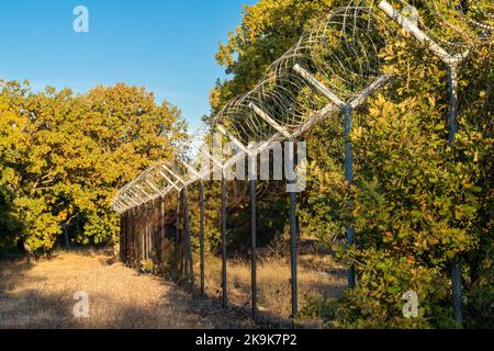 Blick auf einen hohen Zaun mit Stacheldraht, der durch den Wald an der Grenze zwischen der Türkei und Bulgarien führt Stockfoto