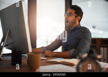 Er zeigt Engagement auch durch die Erschöpfung. Ein junger Geschäftsmann gähnt, während er spät an einem Computer in einem Büro arbeitet. Stockfoto