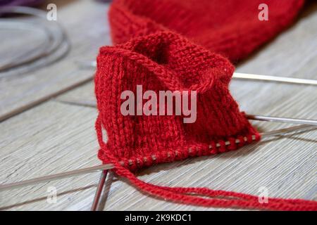 Sock im Prozess des Strickens mit Nadeln und Garn der roten Farbe auf einem grauen Holztisch. Nahaufnahme Stockfoto