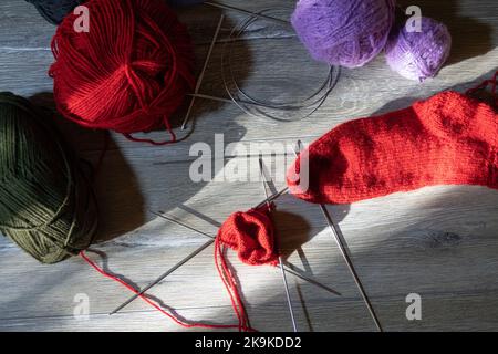 Sock im Prozess des Strickens mit Nadeln und Garn der roten Farbe auf einem grauen Holztisch. Schlint Fäden von rot, lila, dunkelgrün und schwarz auf Stockfoto