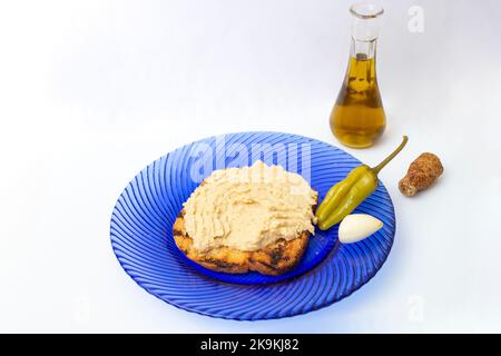 Humuspaste mit Knoblauch, eingelegtem grünem Pfeffer und Sesamöl auf einer gerösteten Brotscheibe, auf einer blauen Glasplatte, isoliert auf weißem Hintergrund. Seite vi Stockfoto