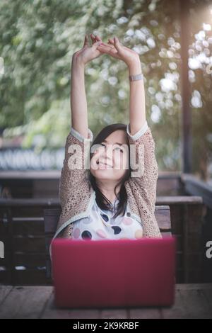 Glückliche asiatische Frau heben den Arm von Körper und Muskeln, während sie mit einem Laptop am Schreibtisch im Home Office arbeitet, lächelnde reife Frau, die sich von com ausruht Stockfoto