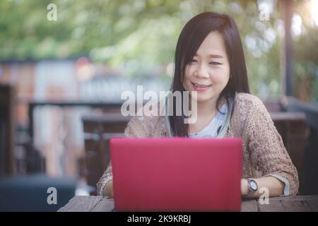 Eine lächelnde asiatische Frau, die bequem online auf ihrem Laptop außerhalb des Büros arbeitet. Konzept der Technik und der modernen Arbeit Stockfoto
