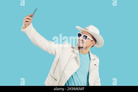 Junger glücklicher kaukasischer Mann in Sonnenbrille nimmt Selfie auf Handy, steht in türkisfarbenem Studio Stockfoto