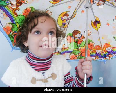 Moskau, Russland, Mai 2011: Niedliches Kleinkind-Mädchen mit riesigen blauen Augen schaut auf, beißt Lippen. Unter dem Regen mit hellem Regenschirm. Stockfoto