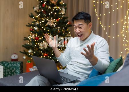 Ein asiatischer Mann liest zu Weihnachten glückliche Nachrichten auf einem Laptop, ein Geschäftsmann, der während der Neujahrsferien auf dem Sofa im Wohnzimmer sitzt und den Siegeserfolg feiert. Stockfoto