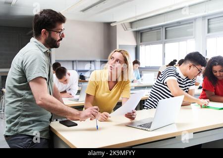 Junger männlicher Lehrer hilft weiblichen Schüler in der Klasse. Der Tutor hilft seinen Schülern, indem er die Lektion erklärt und Zweifel löst. Stockfoto