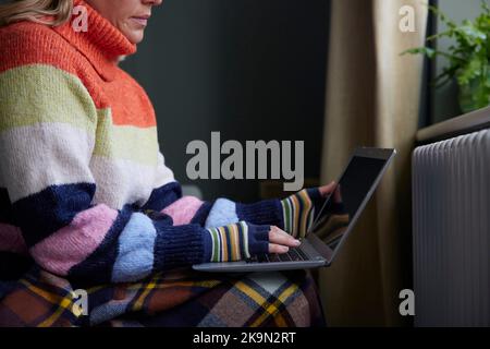 Frau In Handschuhen Mit Laptop Versucht, Durch Heizkörper Während Der Lebenshaltungskosten Energiekrise Warm Zu Halten Stockfoto