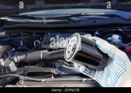 Der Automechaniker hält einen Ölfilter in der Hand, um ihn auszutauschen, und der Motor des Fahrzeugs befindet sich auf einem verschwommenen Hintergrund. Geringer Fokus. Stockfoto