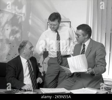 1989, historisch, in einem Büro, versammelten sich drei leitende Führungskräfte eines Unternehmens an einem Schreibtisch, um einen Bericht zu diskutieren, in dem die Gewinnentwicklung und Geschäftsprognosen detailliert beschrieben wurden, England, Großbritannien. Stockfoto