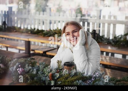 Schöne lächelnde blonde Frau sitzt auf einer Holzbank, träumt, denkt, hält den Kopf mit den Händen in gestrickten Fäustlingen mit Einweg-Pappbecher und Tannenbrause Stockfoto