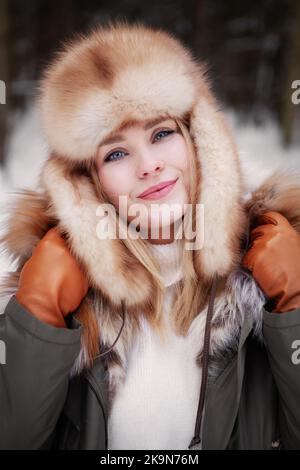 Frau Gesicht und Brust Porträt Nahaufnahme in Jacke, Ohrenklappen Pelzhut und orange Lederhandschuhe, Mädchen mit freundlichen Lächeln, blaue Augen. Schönes kaukasisches fa Stockfoto