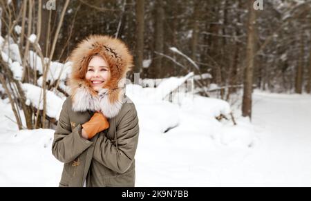 Glückliche Frau lächelt und hüllt sich in warme Kleidung im Winterwald auf Schnee Hintergrund. Tragen stilvolle Mode-Outfit khaki Parka Mantel Jacke mit Stockfoto