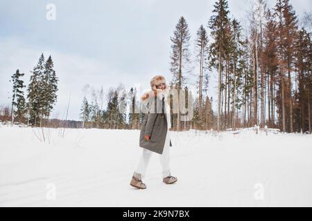 Mädchen zu Fuß im Winter Schneewiese, trägt stilvolle Mode-Outfit khaki Parka Mantel Jacke, Pelzhut und Mond Stiefel, in voller Länge, Blick zurück. Glücklich s Stockfoto
