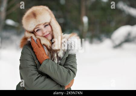 Winterporträt der schönen Frau wickeln und umarmen sich in Pelzjacke, tragen Ohrenklappen Hut und Lederhandschuhe, gemütliches Porträt auf Schnee Hintergrund outd Stockfoto