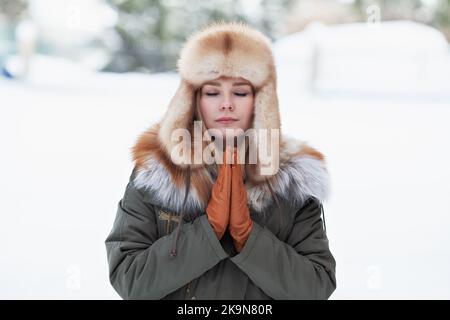 Frau entspannt sich und meditiert im Winter im Freien. Ruhige schöne Frau beten Porträt im Winter Schnee Hintergrund mit Händen in namaste Yoga-Position und c Stockfoto
